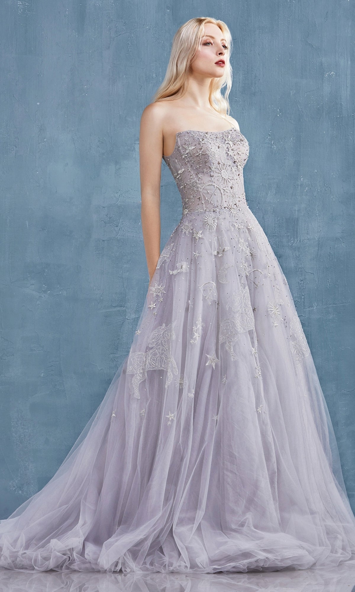 Against a textured blue background, a woman wears the Constellation-Print Strapless Prom Ball Gown A0890—a floor-length lavender dress featuring intricate embroidery and a flowing tulle skirt.